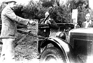 Bob Emslie pointing, an irate Dick Latta with a gun, at the Cudgery gate on the road to Kurnell.  Latta is endeavoring to stop people using the road. Unauthorised vehicles using the road he had personally made were taking little care of their driving techniques and were cutting the road up and not doing anything towards its upkeep. Nor did the Council do anything. The car is a Hupmobile. (Photo: Nola Latta)