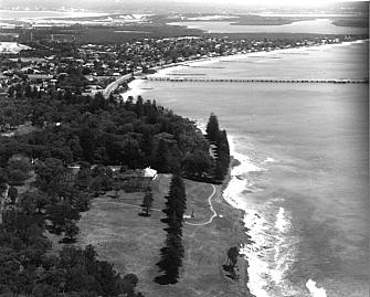Aerial view of Kurnell, 1999. Captain Cook Landing Place Reserve and Alpha House in the foreground, Kurnell Village facing Botany Bay, Towra Point in the upper right. The Reserve wharf, seen in the previous photograph, was demolished by the 1974 storms. Caltex wharf and the sand-stabilising groynes jut out into Botany Bay from Silver Beach.